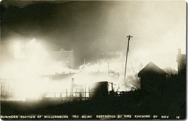 Black and white photograph at night showing several two-story wooden storefronts in flames next to a leaning telephone pole, with smaller black buildings silhouetted in the foreground